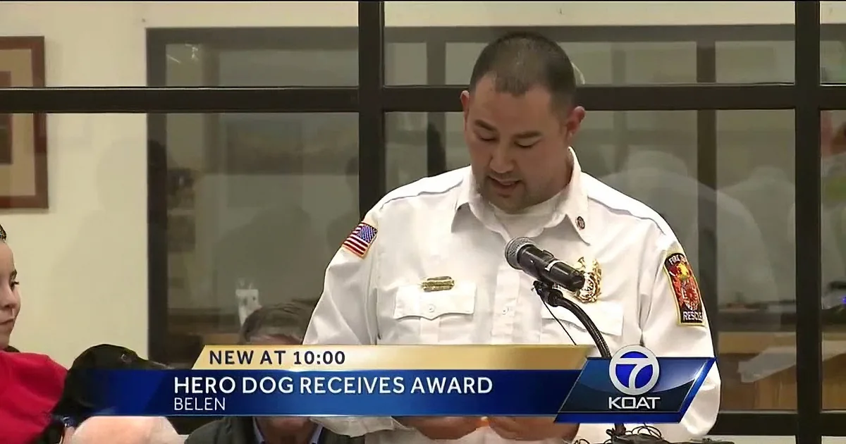 A man in a white uniform speaks at a podium, with a banner below reading "Hero Dog Receives Award."