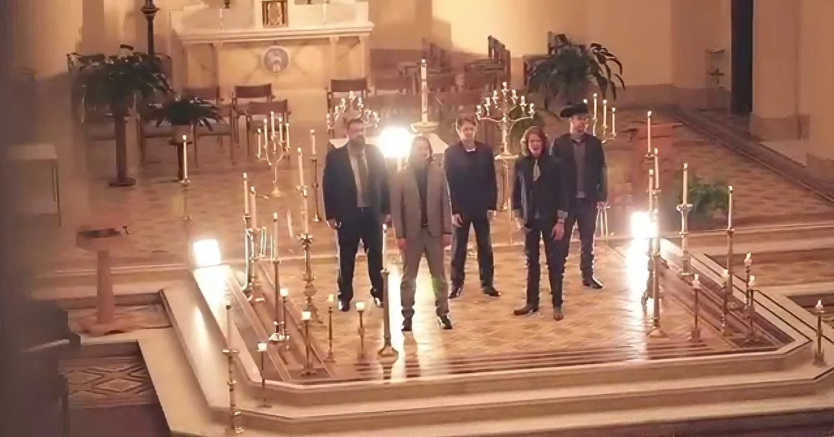 A group of six people stands in a church aisle, surrounded by lit candles and ornate decor.
