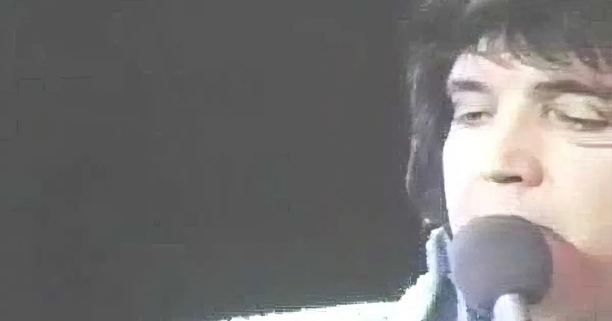 A person holding a microphone, possibly singing or speaking, with a dark background.