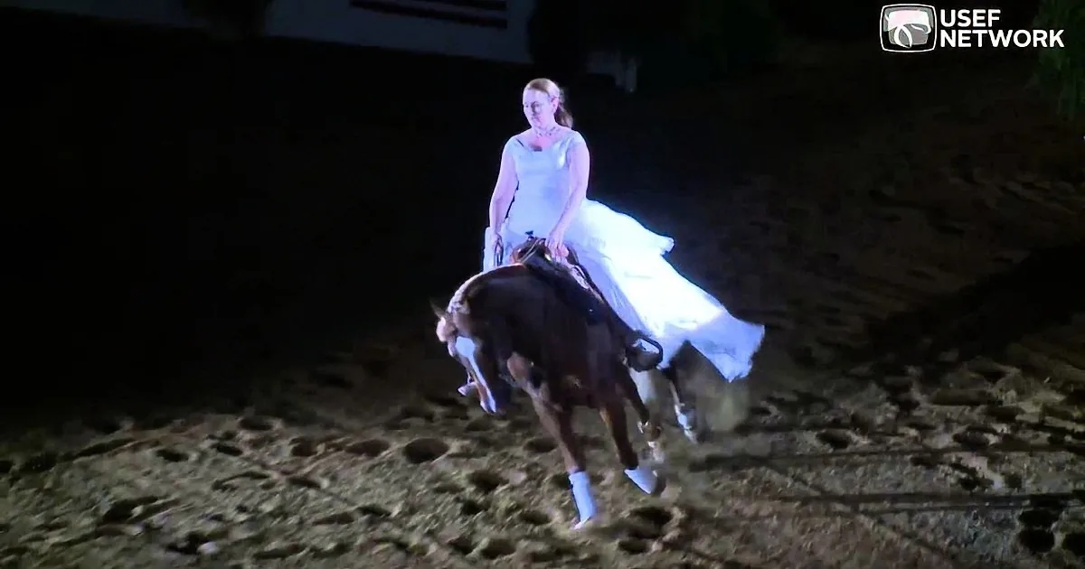 A woman in a flowing white dress rides a horse on a sandy arena at night.