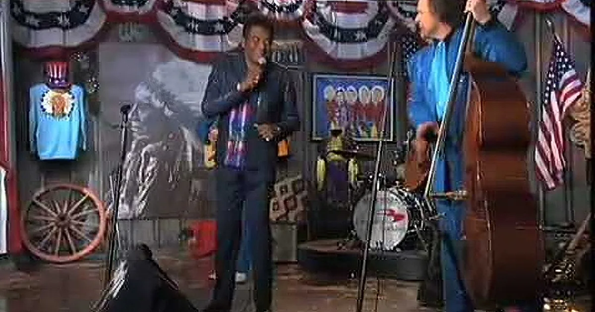 A man in a striped shirt and suit stands on stage, holding a microphone, with a band and patriotic decorations behind him.