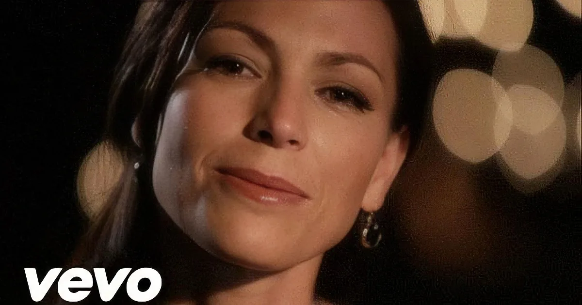 A woman with dark hair and makeup, wearing earrings, is shown in a close-up with blurred lights in the background.