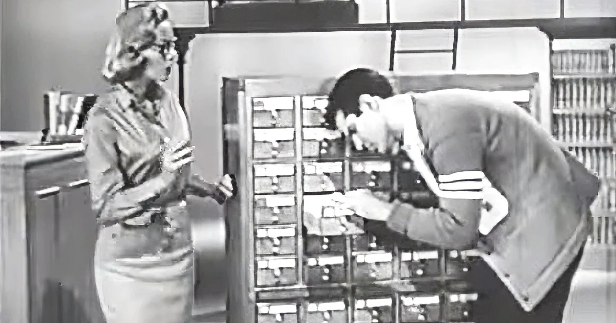 A man searches a card catalog while a woman observes in a library setting.