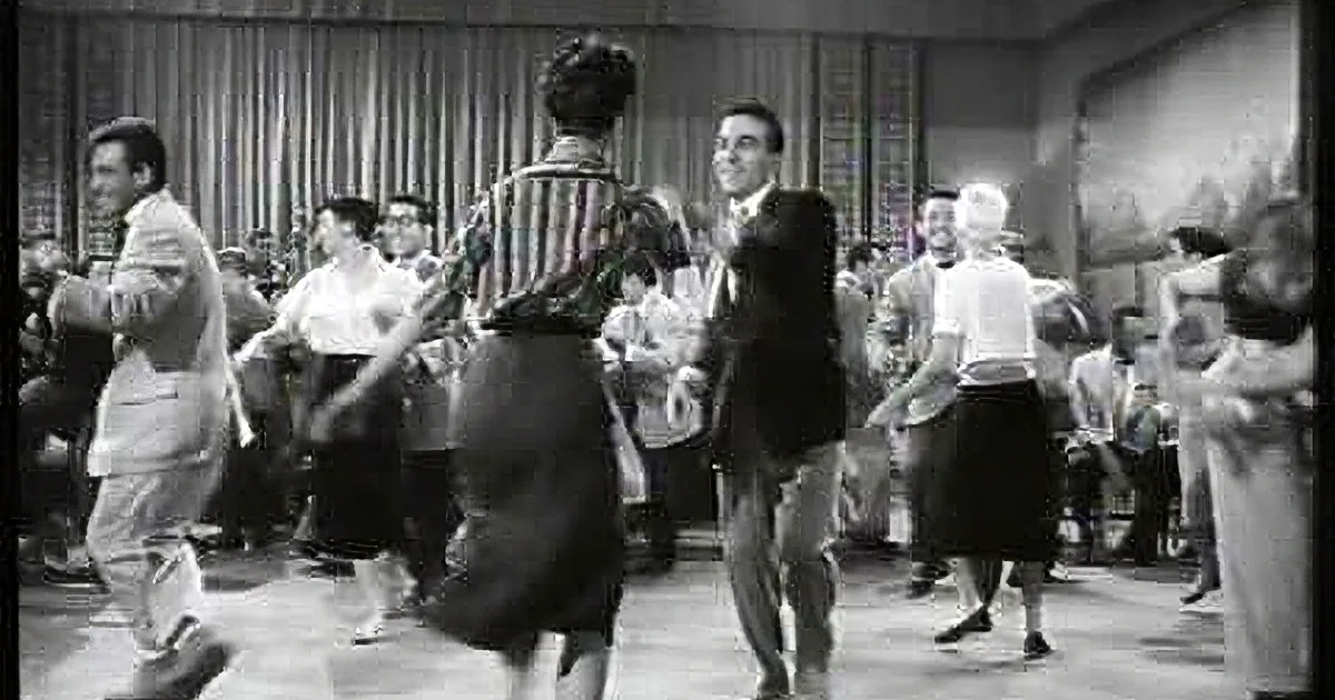 A man in a suit dances energetically with a woman in a black dress, surrounded by a lively crowd in a dance hall.