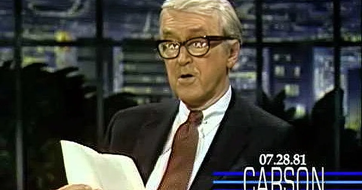A man in a suit and glasses holds a paper, appearing to speak on a news set.