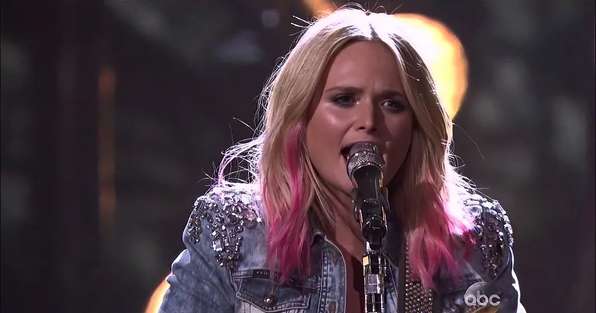 A person with pink-tipped hair sings into a microphone, wearing a denim jacket with studs.