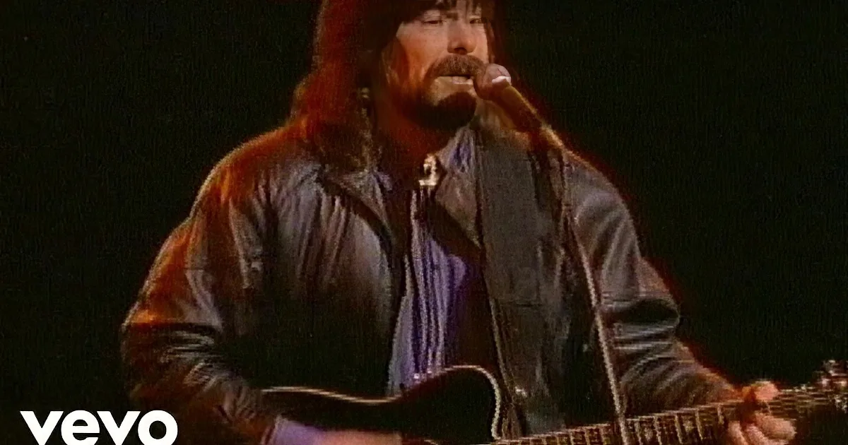 A man with long hair and a beard sings into a microphone while playing an acoustic guitar.