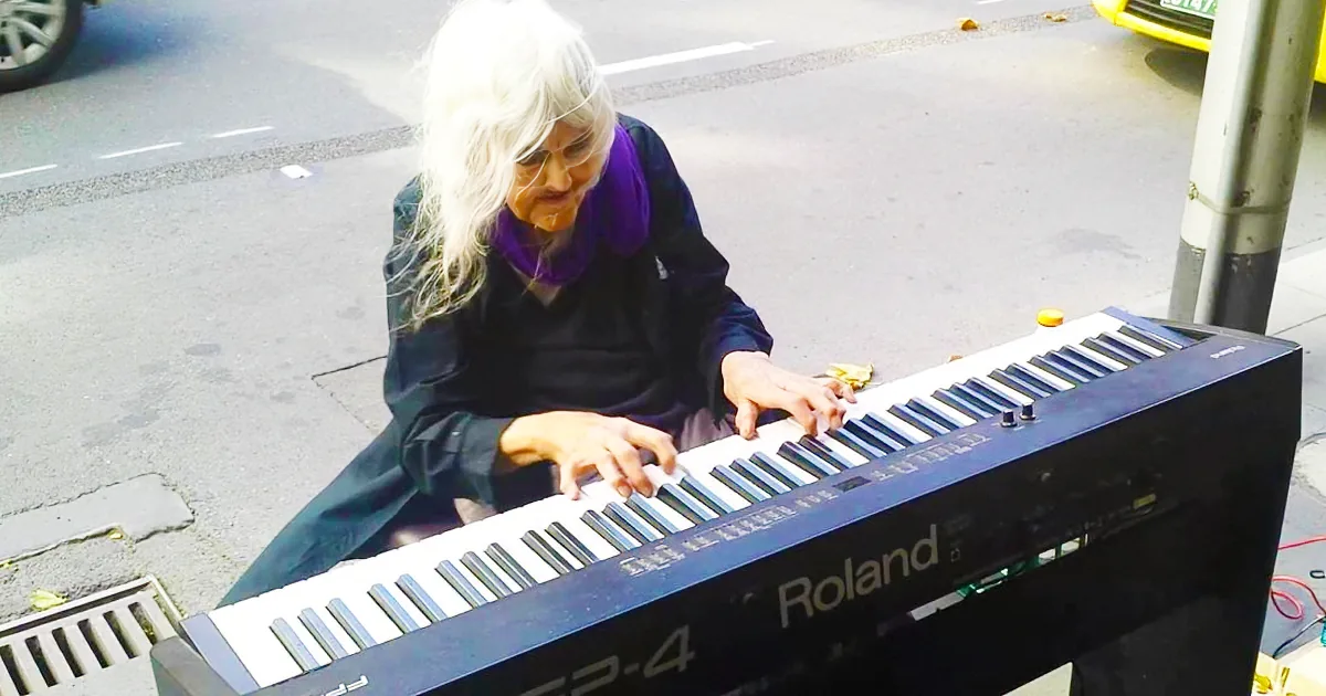 An elderly person with white hair plays a Roland keyboard on a city street.