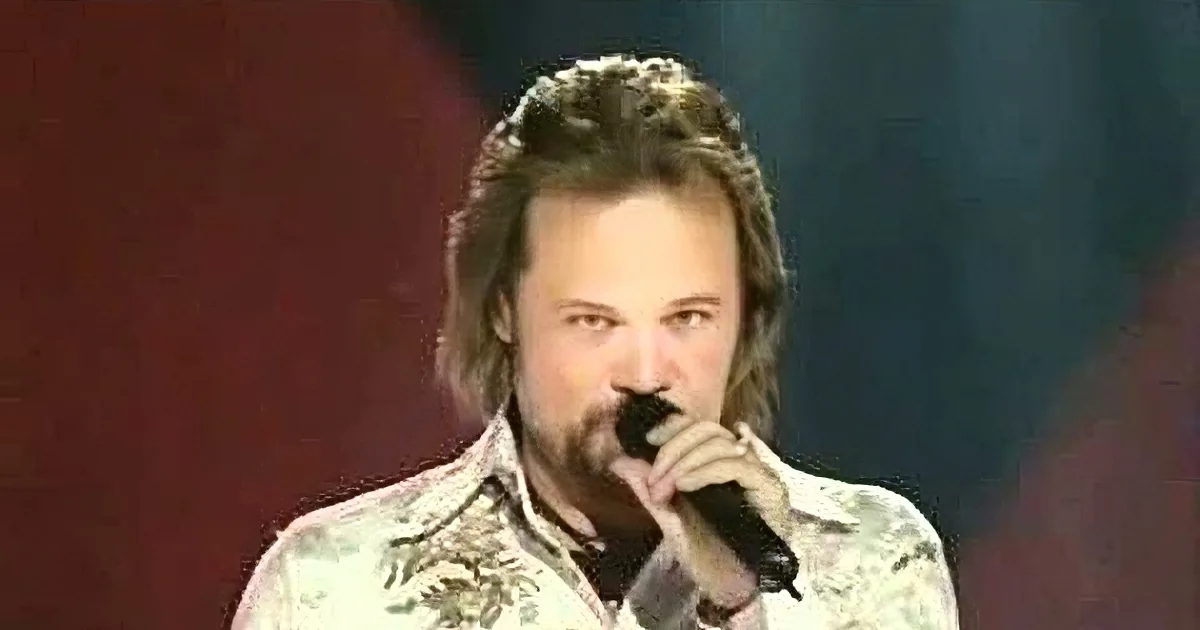 A man with long hair and a beard sings into a microphone, wearing a patterned shirt.
