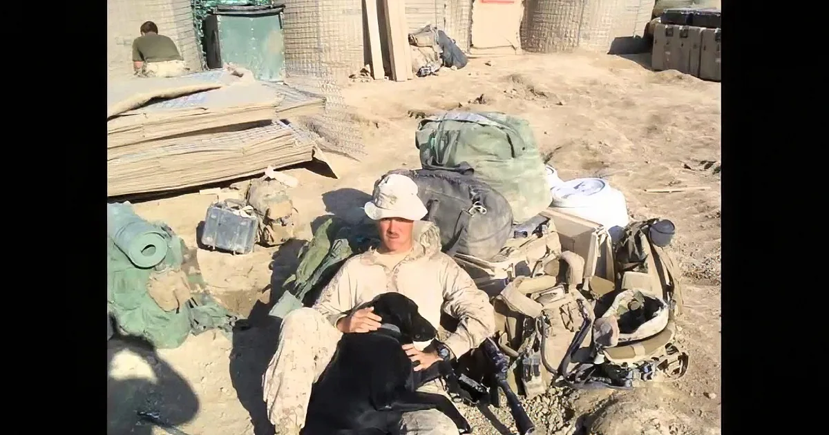 A soldier in a desert uniform sits amidst military gear and equipment on a sandy ground.