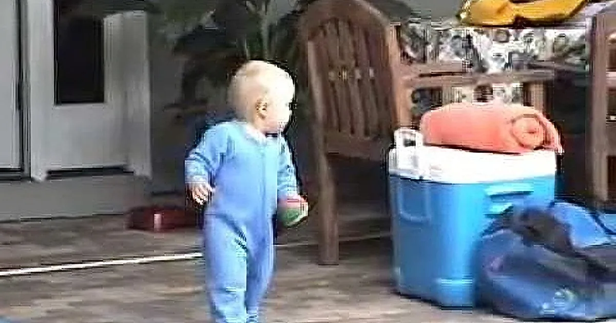 A baby in blue pajamas walks outside near a cooler and chair.