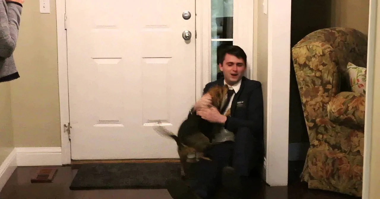 A man in a suit holds a dog, sitting on a step near a door, with a floral chair and another person partially visible.