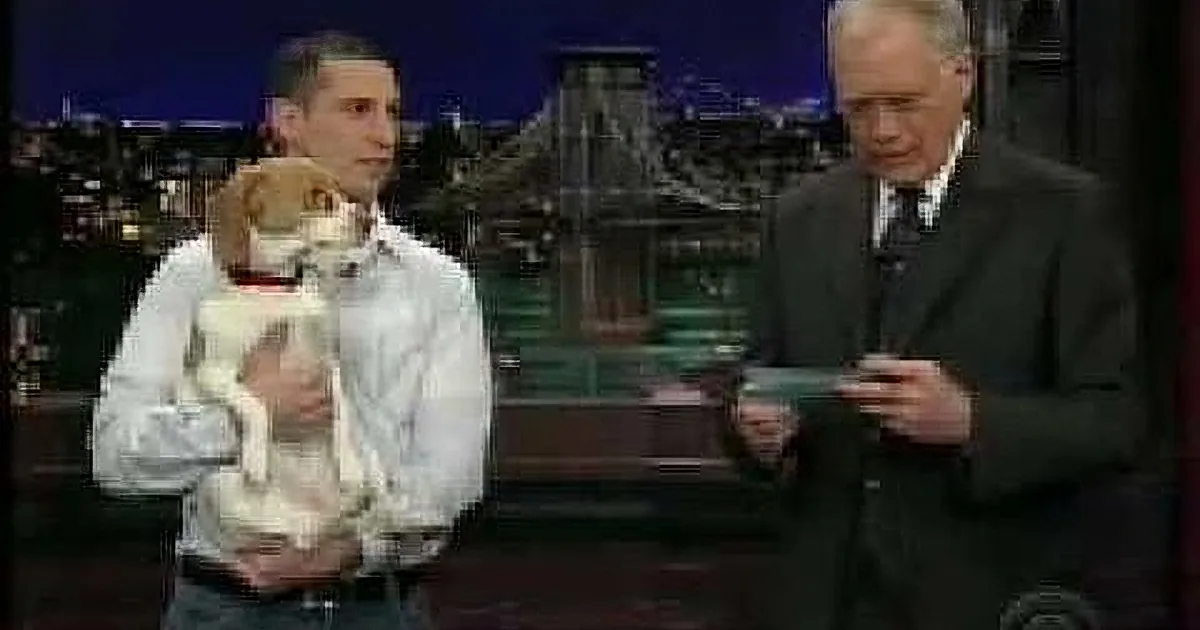 A man holds a small dog while another man in a suit gestures with a drink, both on a talk show set.
