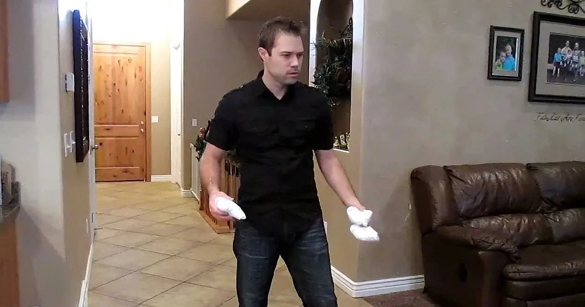A man in a black shirt and jeans holds a white object, possibly a remote, in a living room setting.