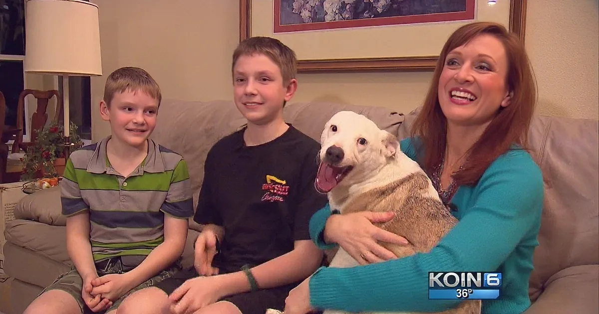 A woman and two boys sit on a couch with a dog, smiling at the camera.