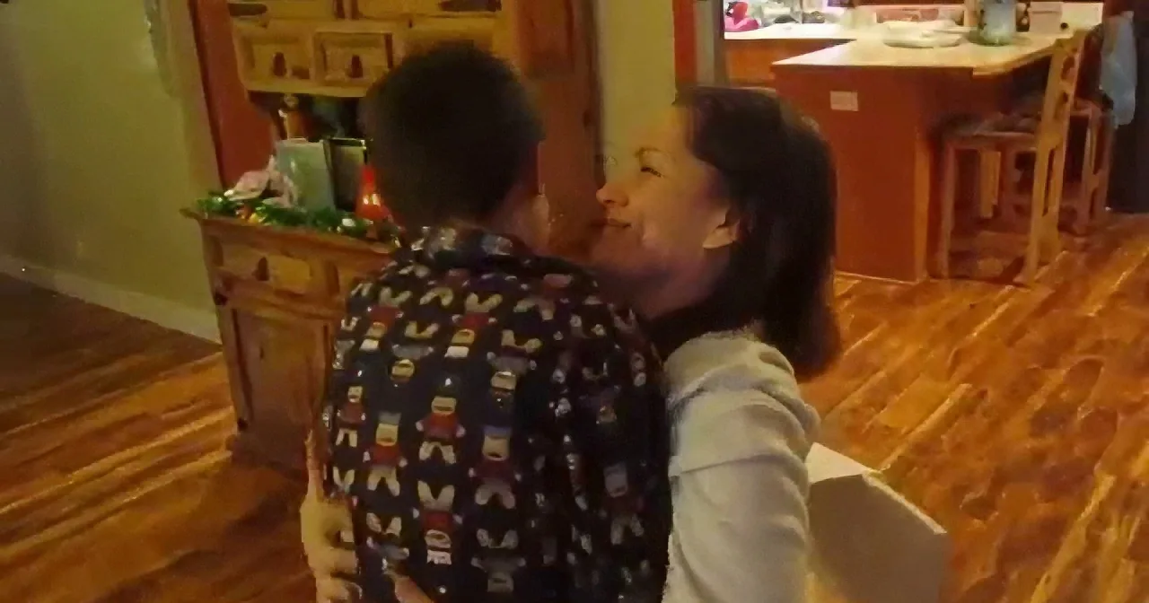 A person in a patterned shirt is being held by another person in a white hoodie in a warmly lit room with wooden floors...