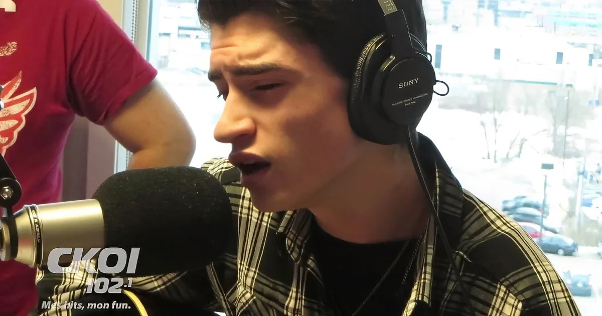 A man wearing headphones sings into a microphone, wearing a plaid shirt, with a window and cityscape in the background.