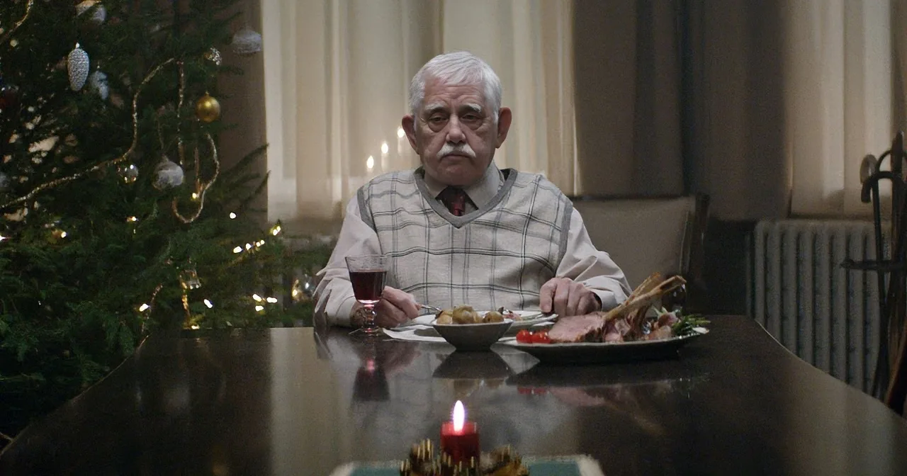An elderly man sits at a table with a Christmas tree and candles, surrounded by food.