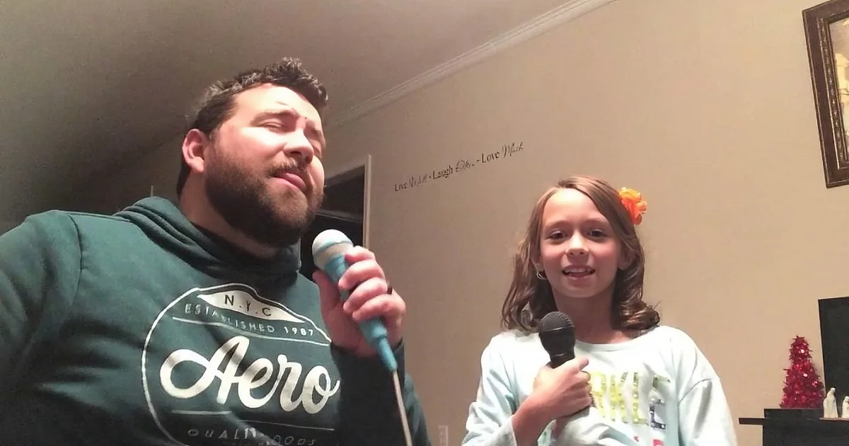 A man and a girl hold microphones, seemingly singing or speaking, in a cozy room.