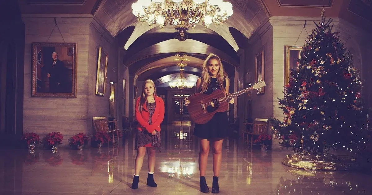 A woman holds a guitar in a grand hall with a Christmas tree, ornate chandelier, and portraits.
