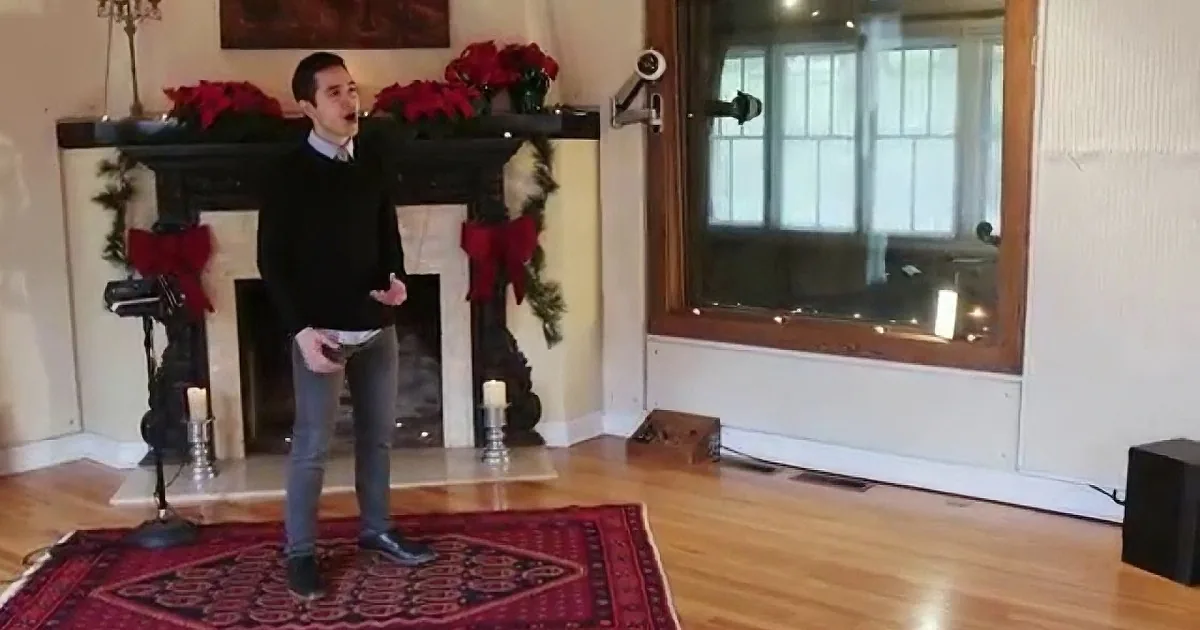 A man stands in a festive living room with a fireplace, red rug, and Christmas decorations.