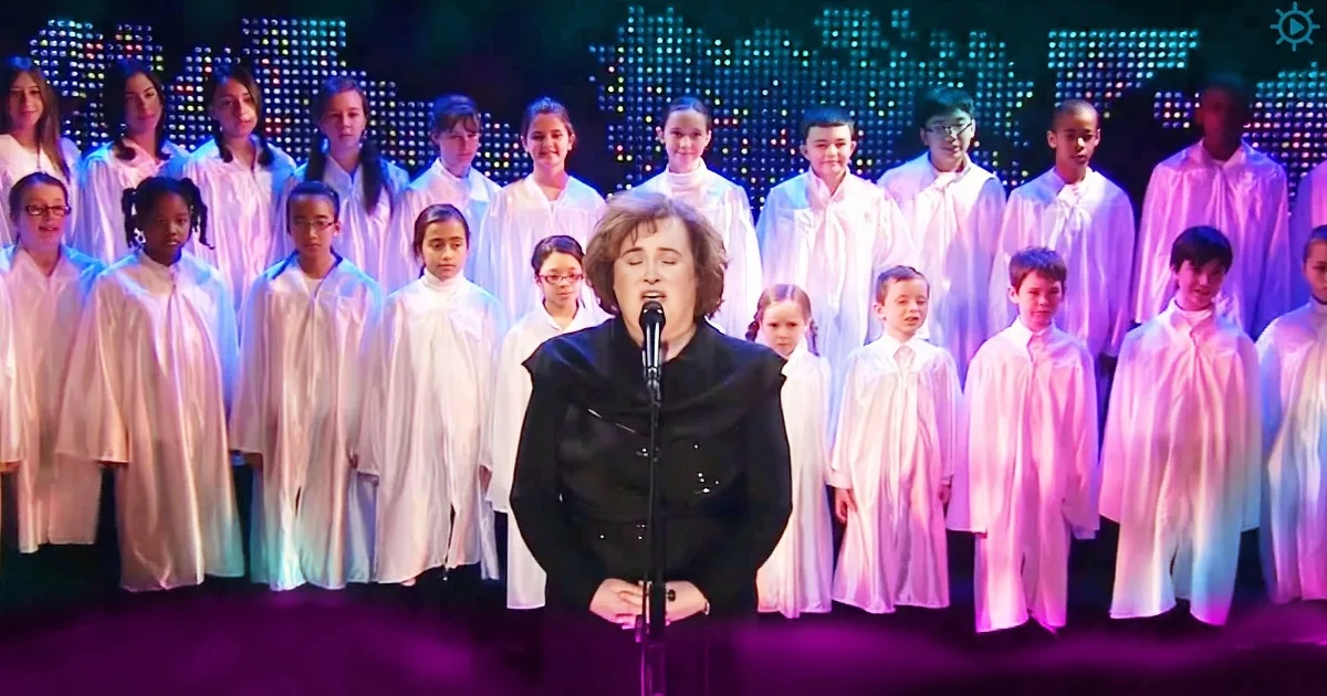 A woman sings into a microphone in front of a choir in white robes under colorful stage lights.