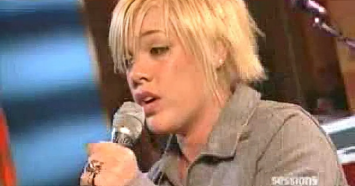 A person with short blonde hair holds a microphone, wearing a brown shirt, in a close-up shot.