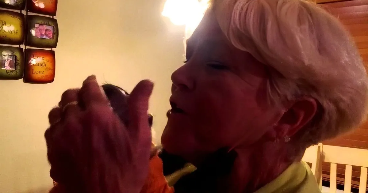 An elderly woman with short gray hair is holding and examining a small object, possibly a watch or a piece of jewelry.