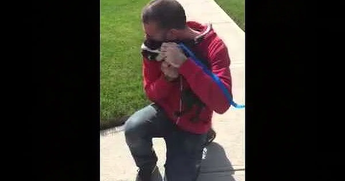A person in a red jacket and jeans is kneeling on a sidewalk, holding a camera and a blue strap.