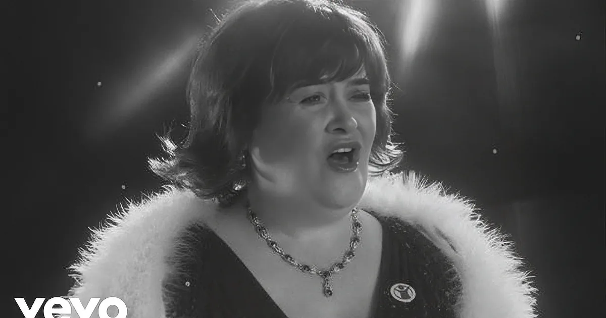 A woman in a fur-trimmed outfit sings passionately, adorned with a necklace and a brooch.