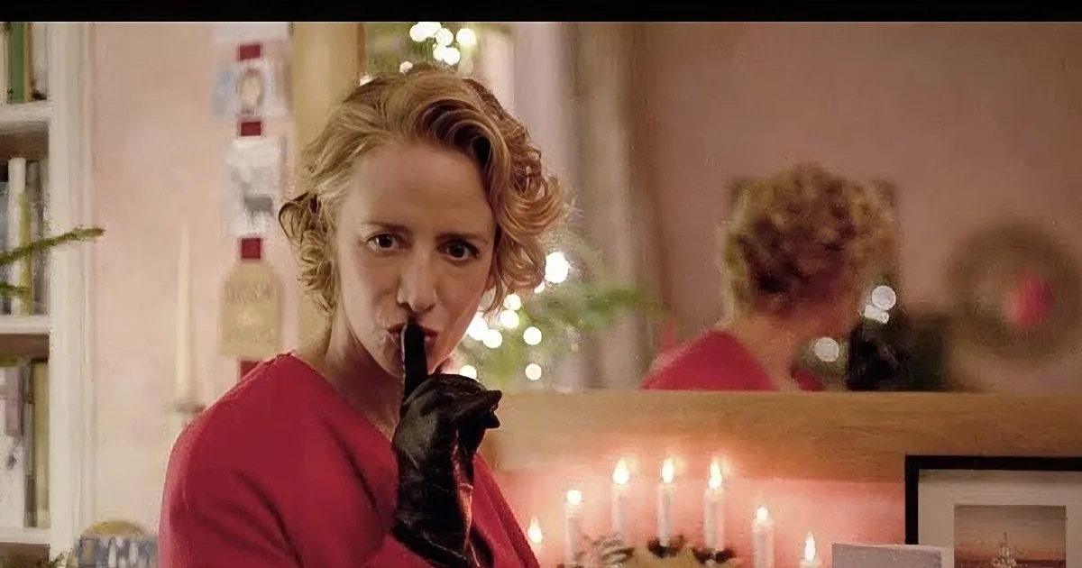 A woman in a red dress and black gloves holds her finger to her lips, surrounded by candles and festive decorations.