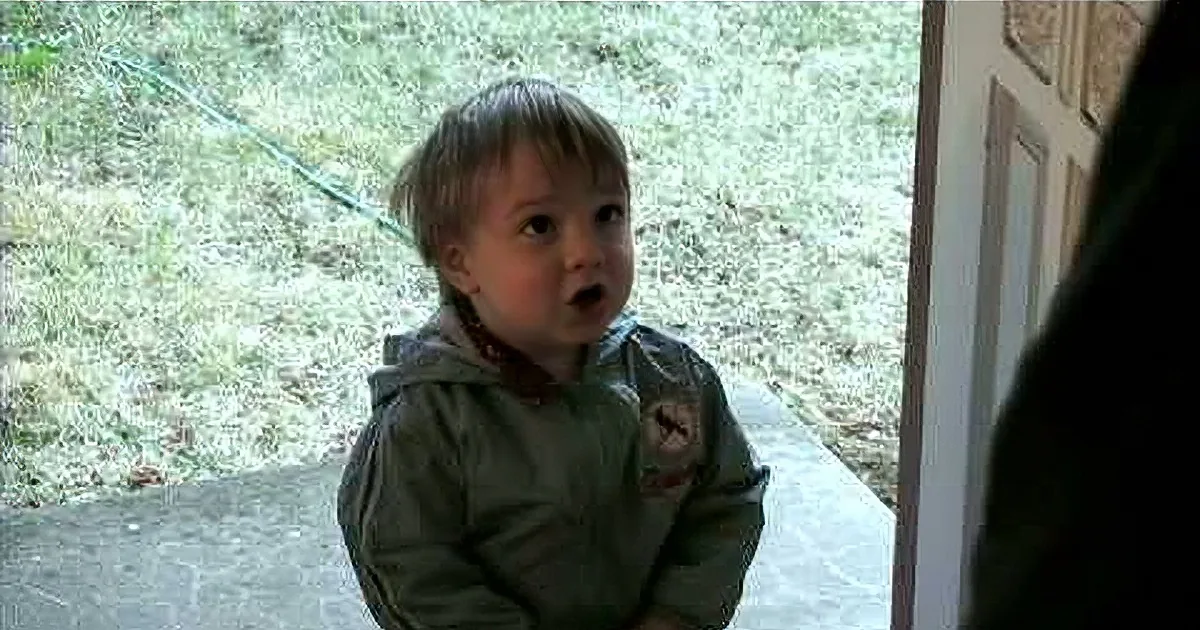 A young child in a green jacket stands on a stone step, looking up with a surprised expression.