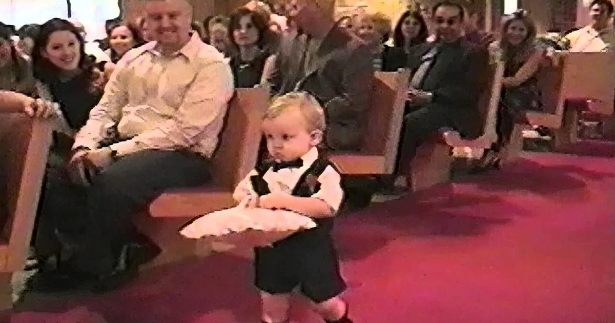 A young child in a tuxedo sits on a red carpet, surrounded by seated adults in a formal setting.