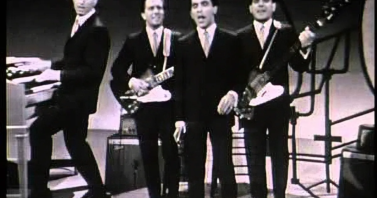 Four musicians in suits perform on stage, one playing a keyboard, another a guitar, while the others sing.