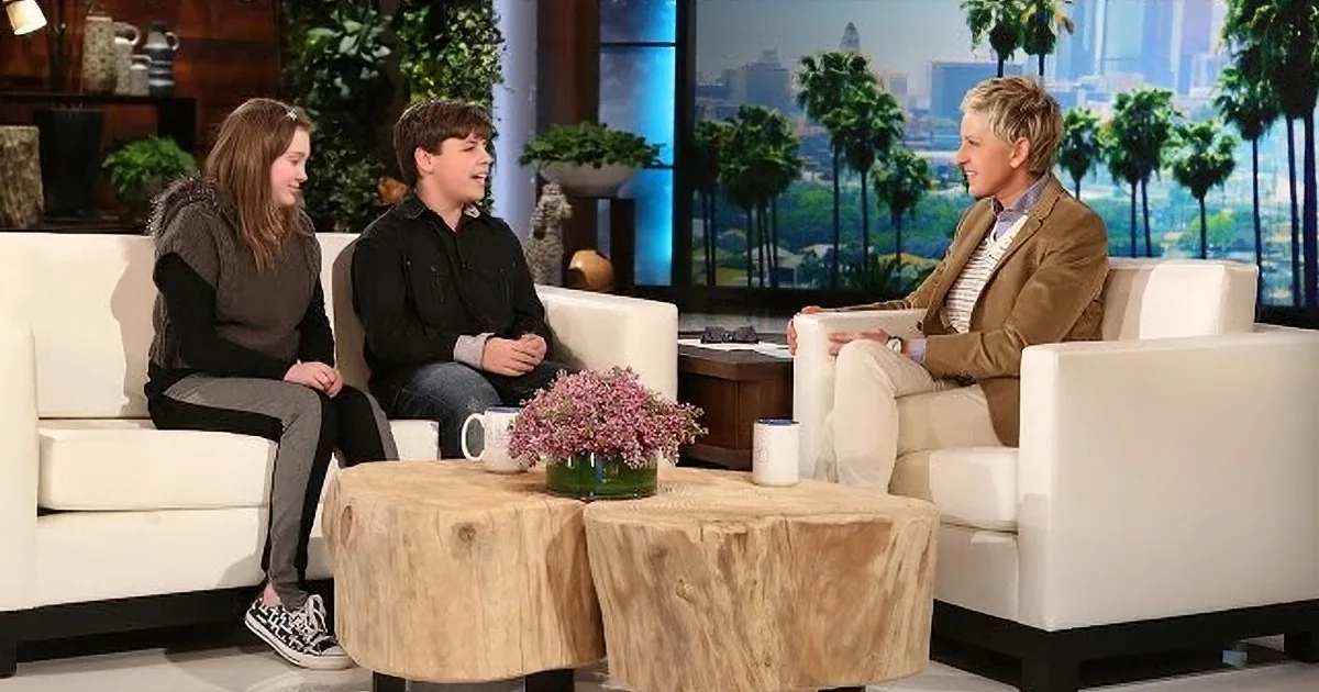 Ellen DeGeneres is seated on a white couch, engaging in conversation with two guests on a talk show set.