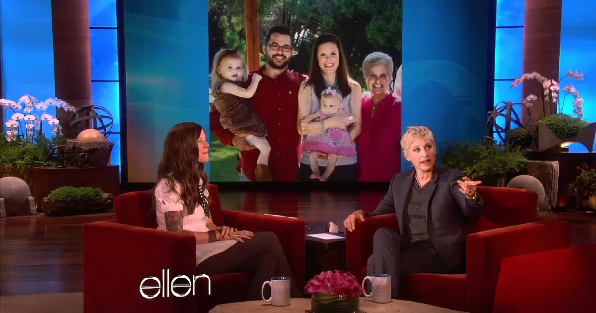 Two women sit on red couches, one holding a baby, discussing on a talk show set with a family photo backdrop.