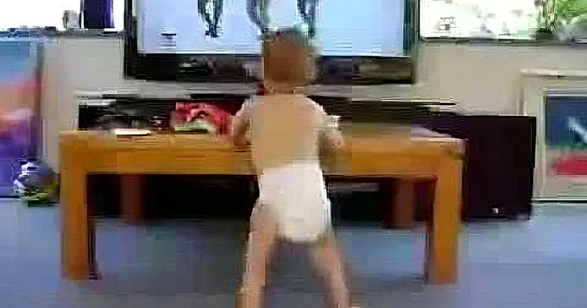 A toddler in a diaper stands at a table, watching a TV showing a dancing figure.