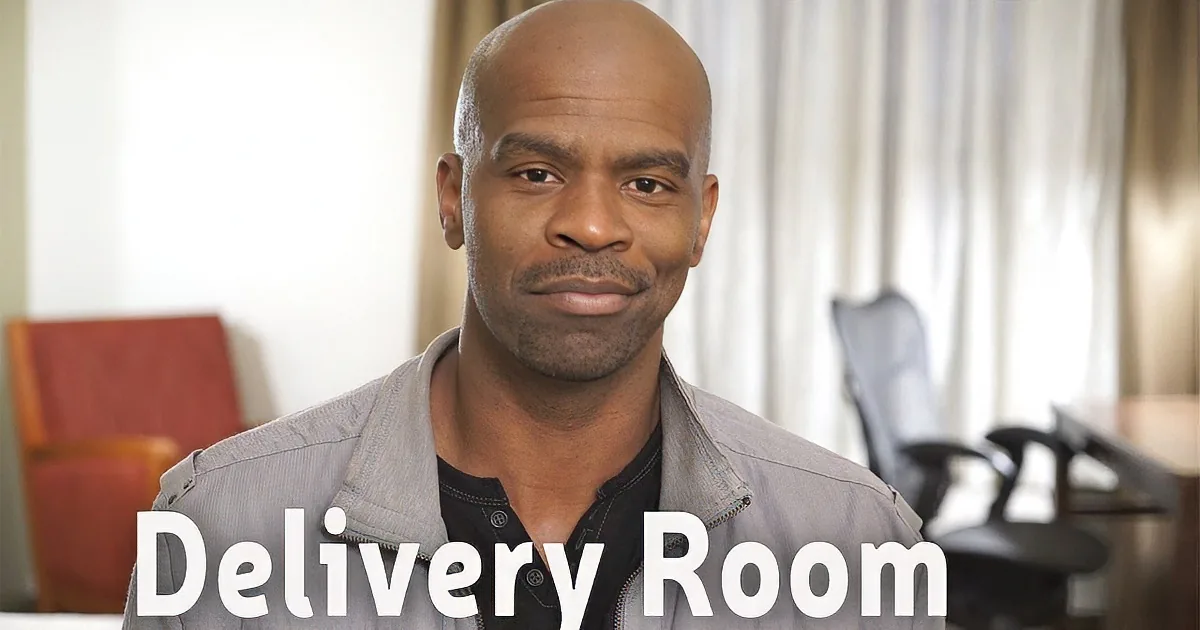 A man with a bald head and a light gray jacket is in a room with chairs and curtains.