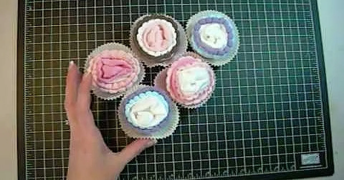 A hand points at six cupcakes with various designs on a cutting mat.