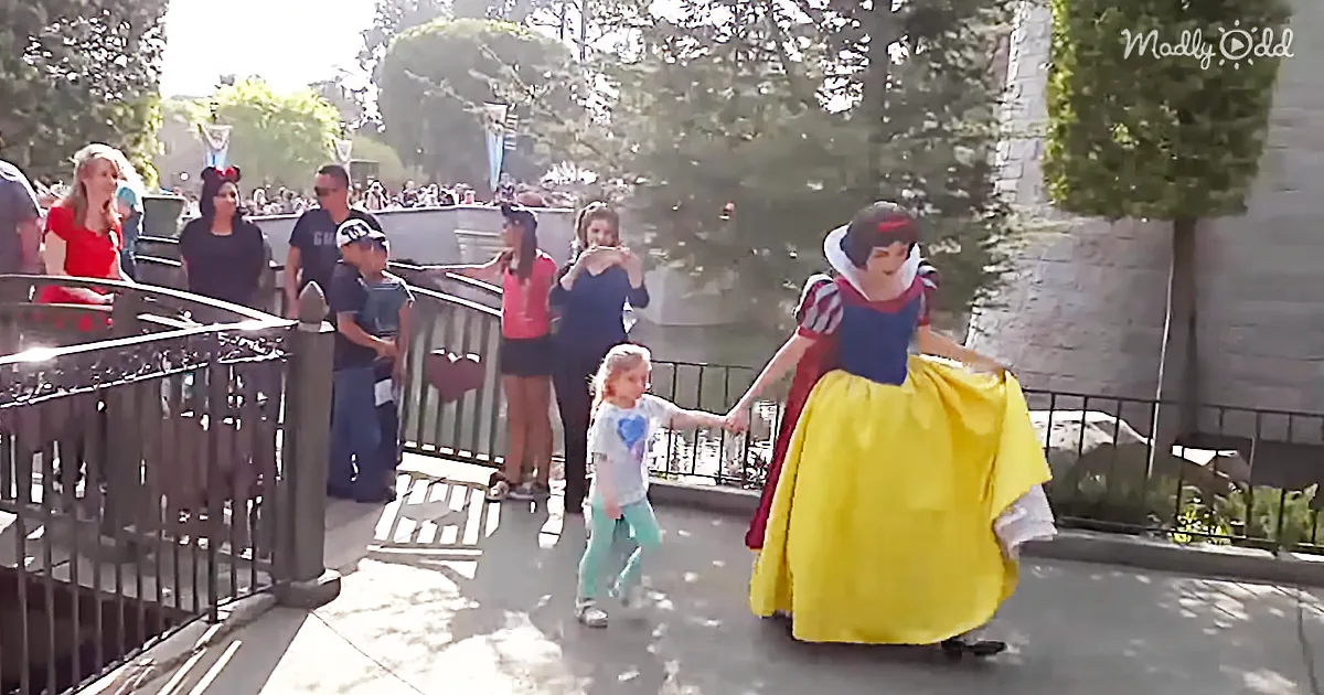 A young girl in a white shirt and blue pants holds hands with a person in a Snow White costume at a Disney park.