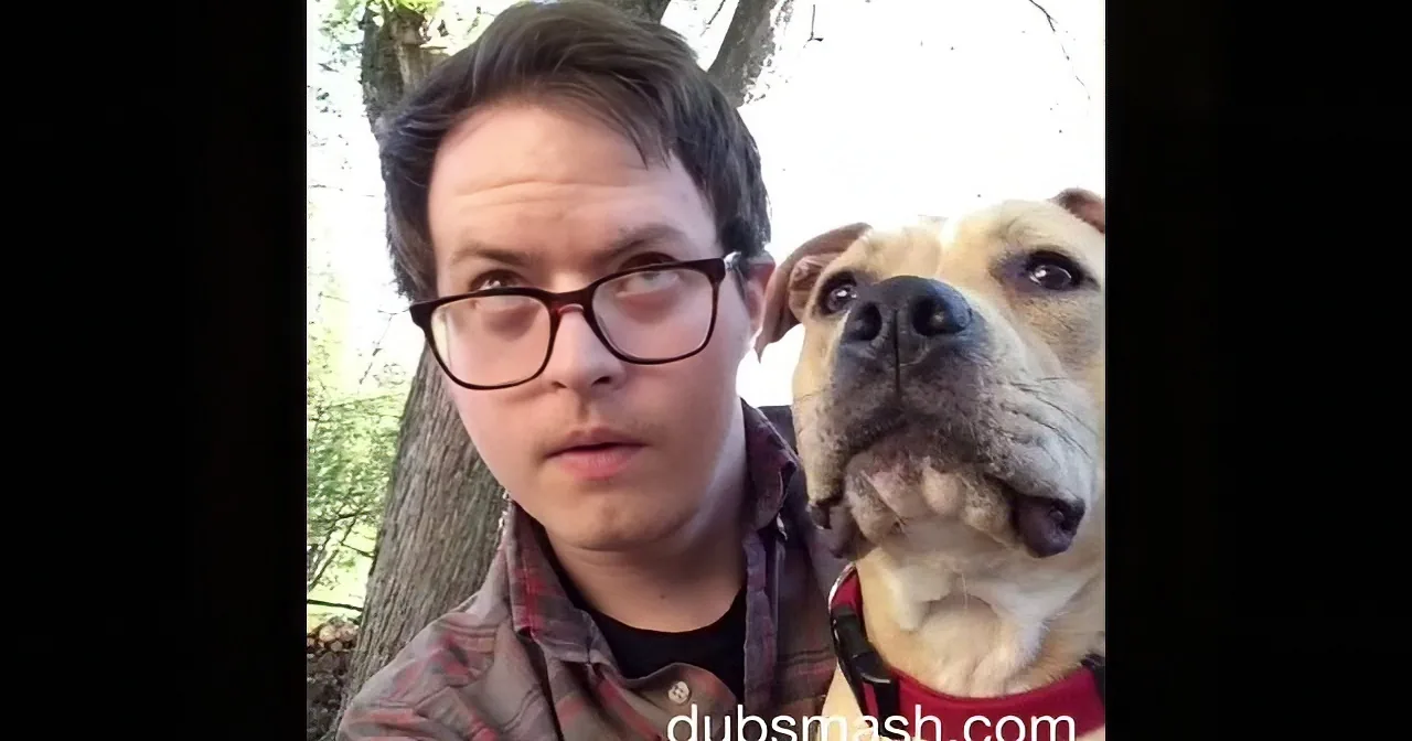 A person and a dog are in a selfie, both looking at the camera with a tree in the background.