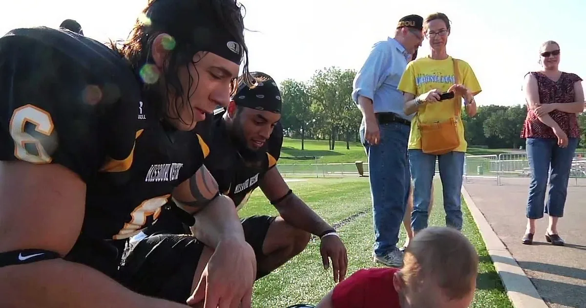 Two football players in black uniforms interact with a child on a grassy field, with spectators in the background.
