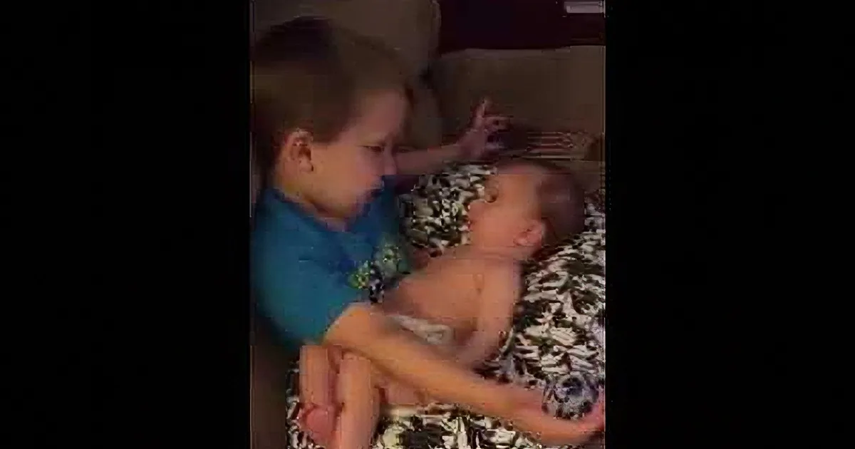 A young boy in a blue shirt is holding and interacting with a baby on a patterned blanket.