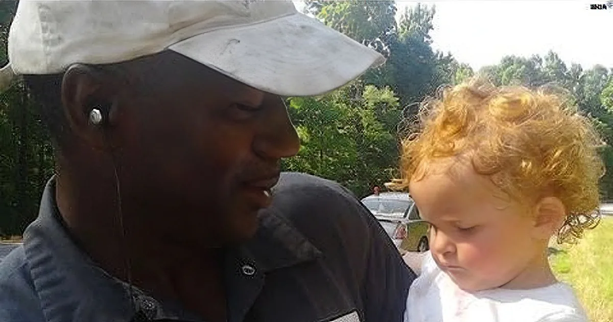A man wearing a cap and earbuds is talking to a young child with curly hair, both outdoors.