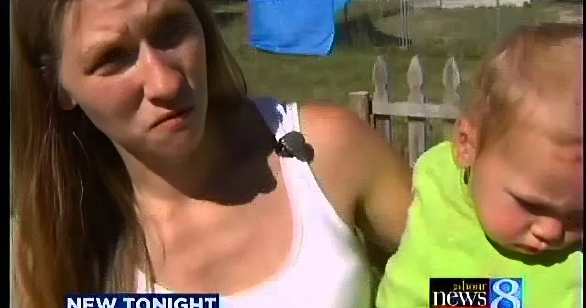 A woman in a white tank top is speaking on a news broadcast, with a baby in a green shirt in the background.