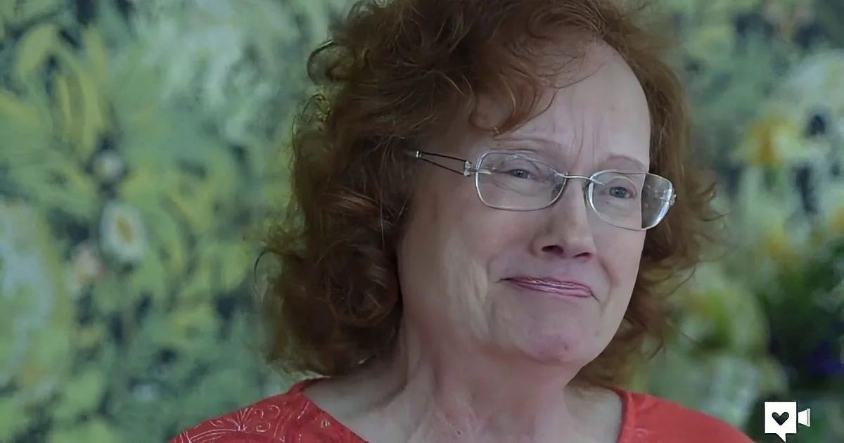 A woman with curly red hair and glasses, wearing a red top, is seen against a blurred green background.
