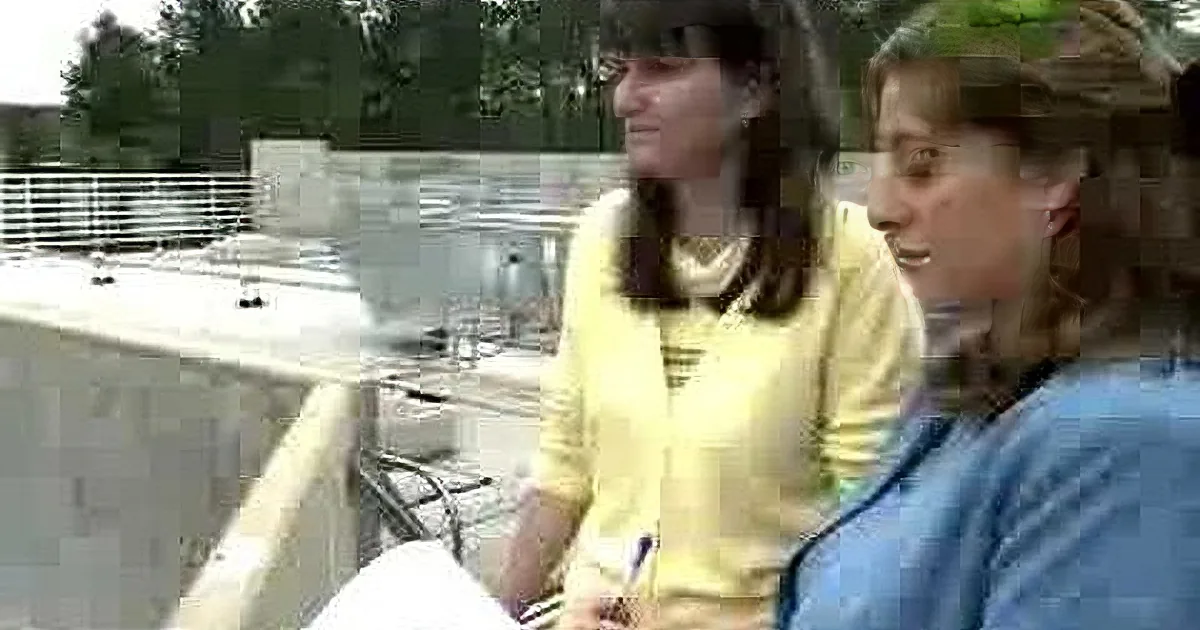 Two women in a conversation, one in yellow, the other in blue, seated outdoors near a railing.