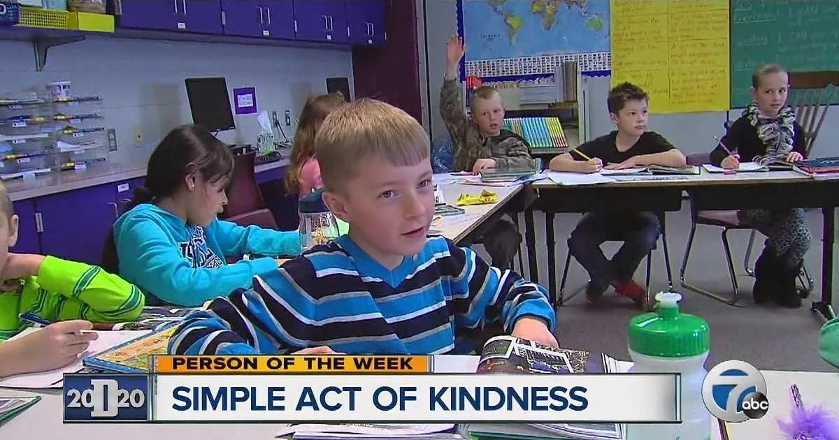 A young boy in a striped shirt sits at a desk in a classroom, surrounded by peers engaged in activities.