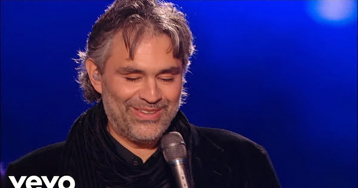 A man with gray hair and beard, wearing a black outfit, sings into a microphone against a blue background.