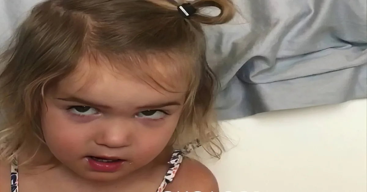 A young child with light brown hair tied in a ponytail, wearing a floral top, looks directly at the camera with a...